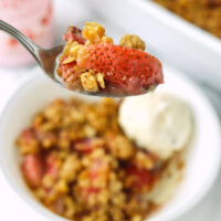Strawberry crisp bite on spoon above bowl. Text overlay "Baileys Strawberries & Cream Strawberry Crisp" and "thatspicychick.com".