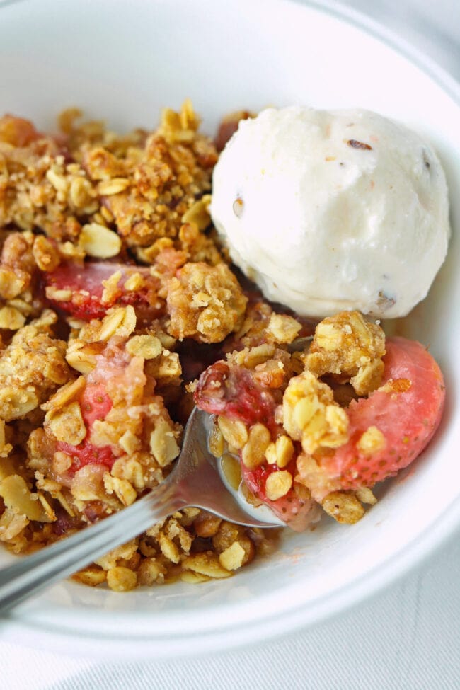 Bowl with a strawberry crisp and ice cream with a bite resting on a spoon.