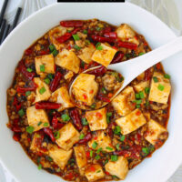 Bowl with spicy tofu and ground pork dish and a spoon, and two rice bowls behind. Text overlay "Braised Tofu & Pork with Pickled Chilies" and "thatspicychick.com".