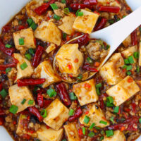 Closeup of spoon in a bowl with spicy tofu and pork dish. Text overlay "Braised Tofu & Pork with Pickled Chilies" and "thatspicychick.com".