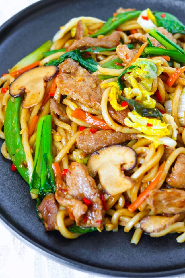 Closeup front view of a black plate with Chinese noodles, pork, and vegetables stir-fry.