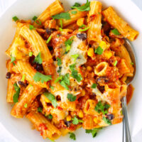 Plate with pasta with corn, black beans, ground chicken, and tomato sauce and a spoon. Text overlay "Chicken Enchilada Pasta", "Cheesy | Spicy | One-Pan Meal" and "thatspicychick.com".