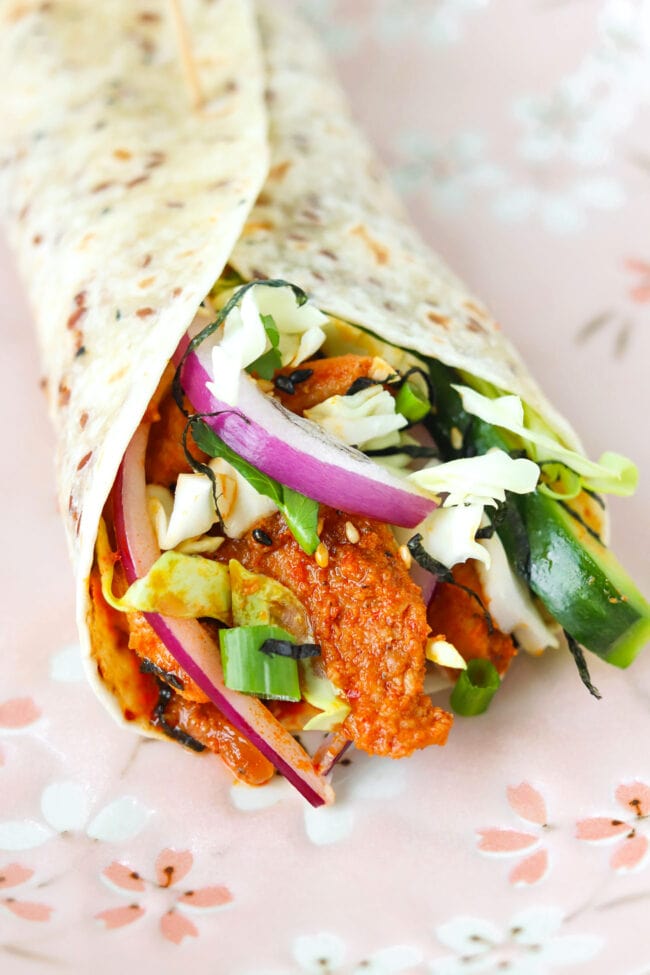 Closeup of gochujang chicken tortilla wrap with veggies, herbs, shredded seaweed, and toasted sesame seeds.