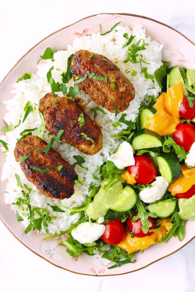 Kofta over steamed rice and salad topped with sauces on a plate.