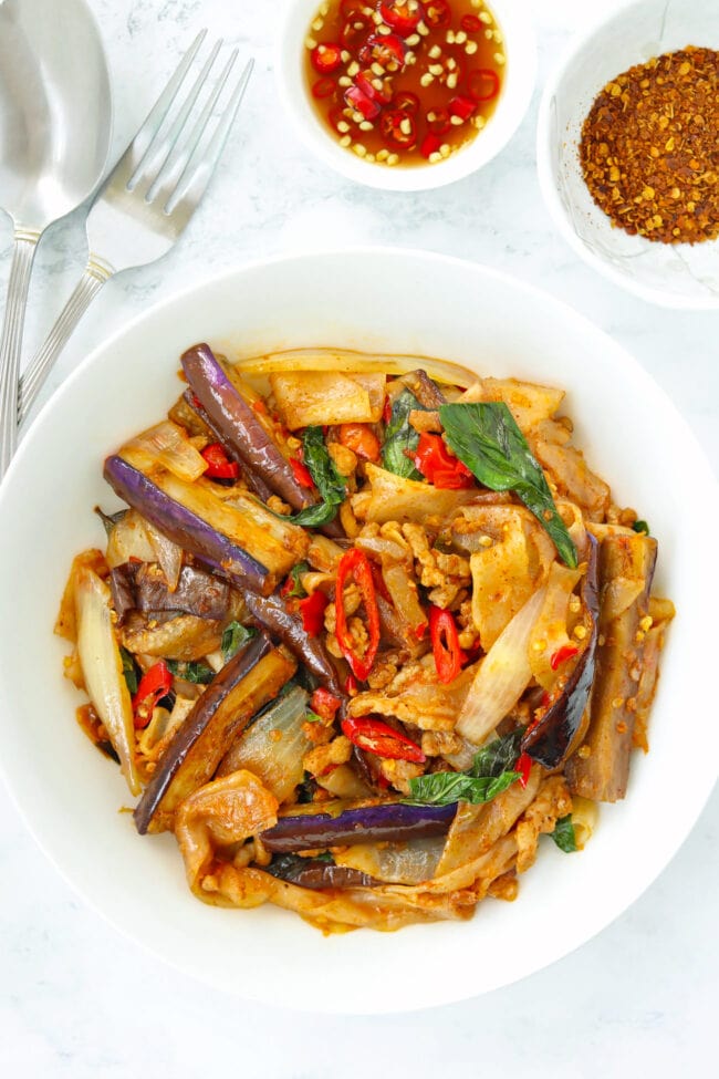 Stir-fried rice noodles with eggplant on a plate. Utensils and condiments in bowls behind.
