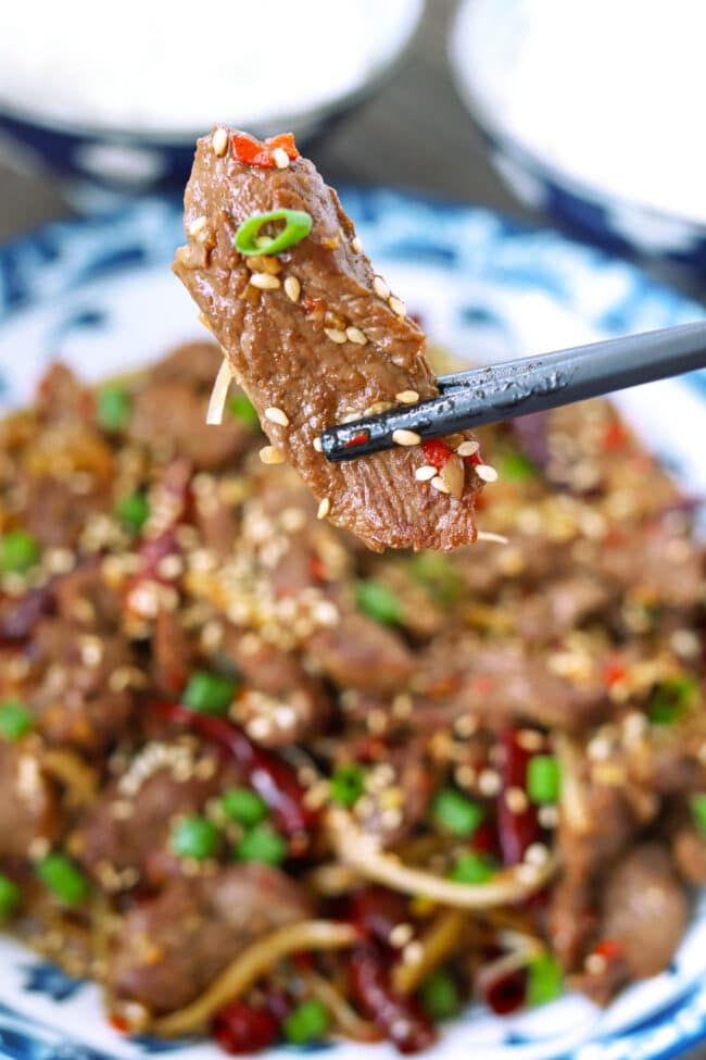 Chopsticks holding up lamb strip above plate with stir-fry.