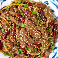 Stir-fried lamb strips garnished with spring onion and sesame seeds on a plate. Text overlay "Mongolian Lamb Stir-fry" and "thatspicychick.com".