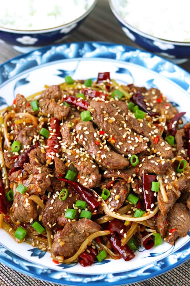 Lamb stir-fry garnished with spring onion and sesame seeds on a plate and two rice bowls.