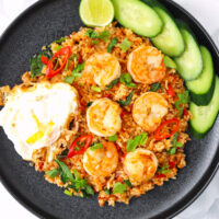 Plate with shrimp fried rice, a fried egg, and cucumber slices and lime wedge. Text overlay "Thai Red Curry Fried Rice" and "thatspicychick.com".