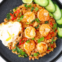Shrimp fried rice on a plate topped with a fried egg and cucumber slices and lime wedge on the side. Text overlay "Thai Red Curry Fried Rice with Shrimp & Thai Style Fried Egg" and "thatspicychick.com".