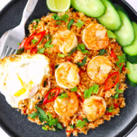 Plate with shrimp fried rice, fork and spoon, fried egg, lime wedge and cucumber slices.