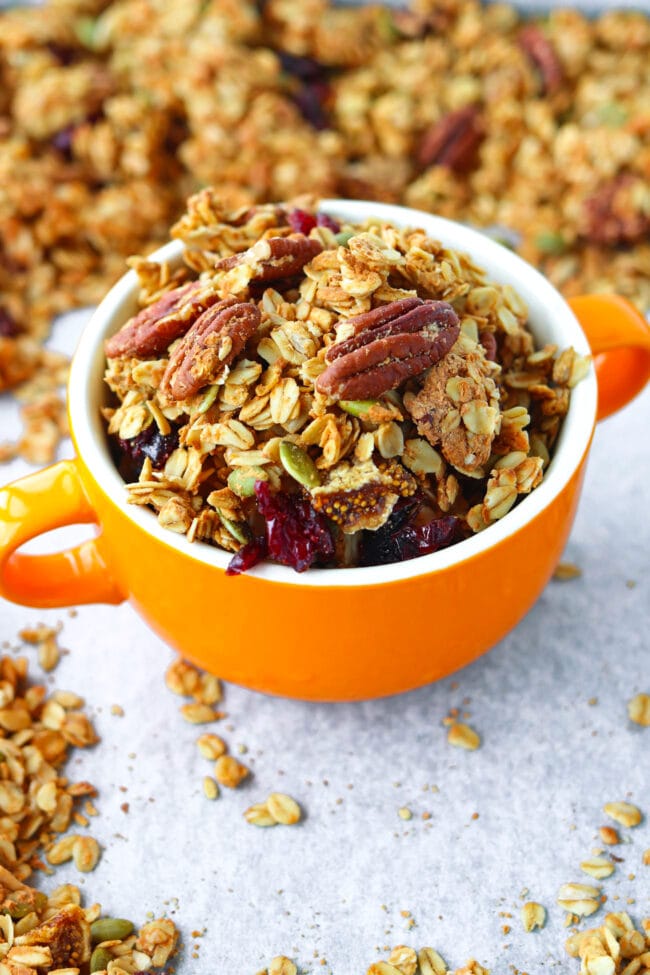 Granola in a bowl with handles in baking tray with scattered granola clusters.