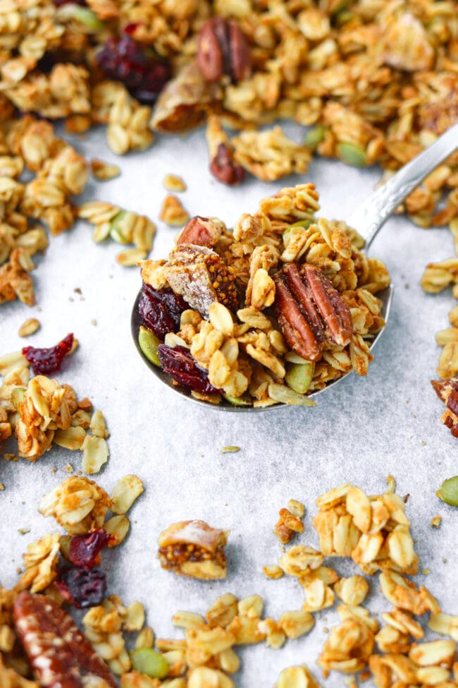 Front view of spoon with granola on a baking tray with scattered granola clusters.