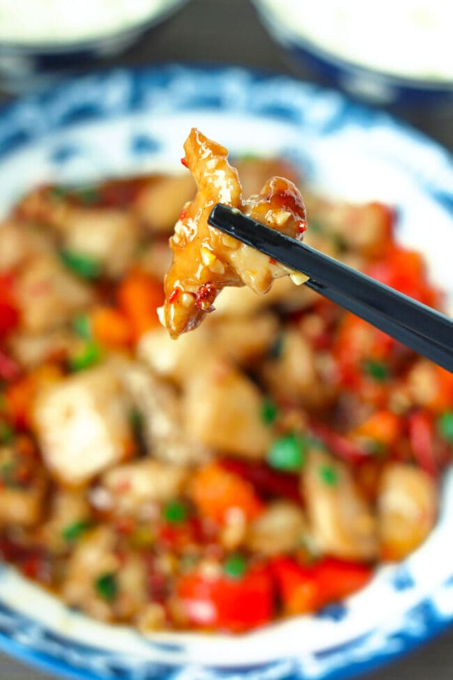 Chopsticks holding up a piece of chicken above a plate with Sichuan Garlic Chicken Stir-fry.