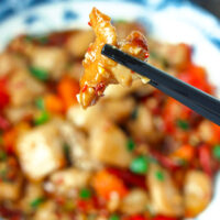 Chopsticks holding up a piece of chicken. Text overlay "Sichuan Garlic Chicken Stir-fry" and "thatspicychick.com".