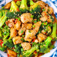 Chicken and broccoli stir-fry closeup top view on a plate. Text overlay "Chicken & Broccoli in Chili Garlic Sauce" and "thatspicychick.com".