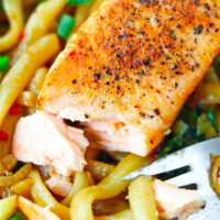 Closeup of salmon fillet flaked with a fork to show inside on udon noodles on a plate. Text overlay "Spicy Cumin Noodles with Salmon" and "thatspicychick.com".