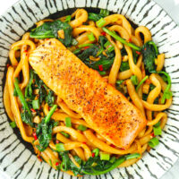 Plate with stir-fried noodles and pan-fried salmon fillet. Text overlay "Spicy Cumin Noodles with Salmon" and "thatspicychick.com".