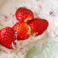 Closeup of bowl with strawberries and bites eaten of pink overnight oats. Text overlay "Strawberry Protein Overnight Oats" and "thatspicychick.com".