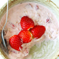 Spoon in bowl with pink overnight oats topped with strawberries. Text overlay "Strawberry Protein Overnight Oats" and "thatspicychick.com".