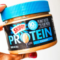 Hand holding up a jar of Skippy creamy protein peanut butter.