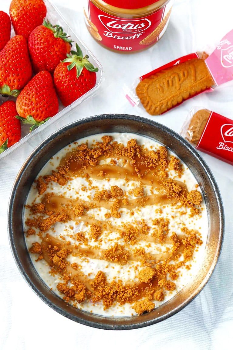 Bowl with Biscoff overnight oats with crumbled Biscoff cookie. Biscoff spread jar, strawberries and cookies behind.
