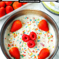 Overnight oats in a bowl with sprinkles and berries. Protein powder tub and strawberries behind. Text overlay "Birthday Cake Overnight Protein Oats" and "thatspicychick.com"