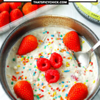 Overnight oats in a bowl with sprinkles, berries and a spoon. Text overlay "Birthday Cake Overnight Protein Oats" and "thatspicychick.com"