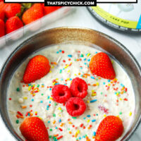 Overnight oats in a bowl with sprinkles and berries. Strawberries and protein powder tub behind. Text overlay "Birthday Cake Overnight Protein Oats" and "thatspicychick.com"