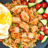 Closeup of fried rice on a plate with a fried egg, tomatoes and cucumber. Text overlay "Chicken Nasi Goreng Indonesian Fried Rice" and "thatspicychick.com".