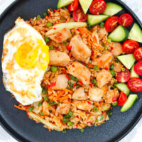Black plate with chicken nasi goreng with a fried egg, sliced cucumber and cherry tomatoes.