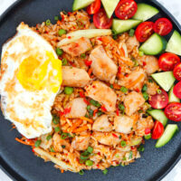 Chicken fried rice on a black plate with tomatoes, cucumber and a fried egg. Text overlay "Chicken Nasi Goreng Indonesian Fried Rice" and "thatspicychick.com".
