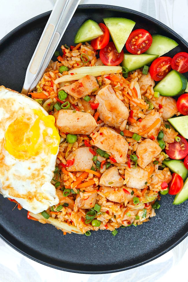 Plate with chicken nasi goreng, fried egg, sliced cucumber and cherry tomatoes and utensils.