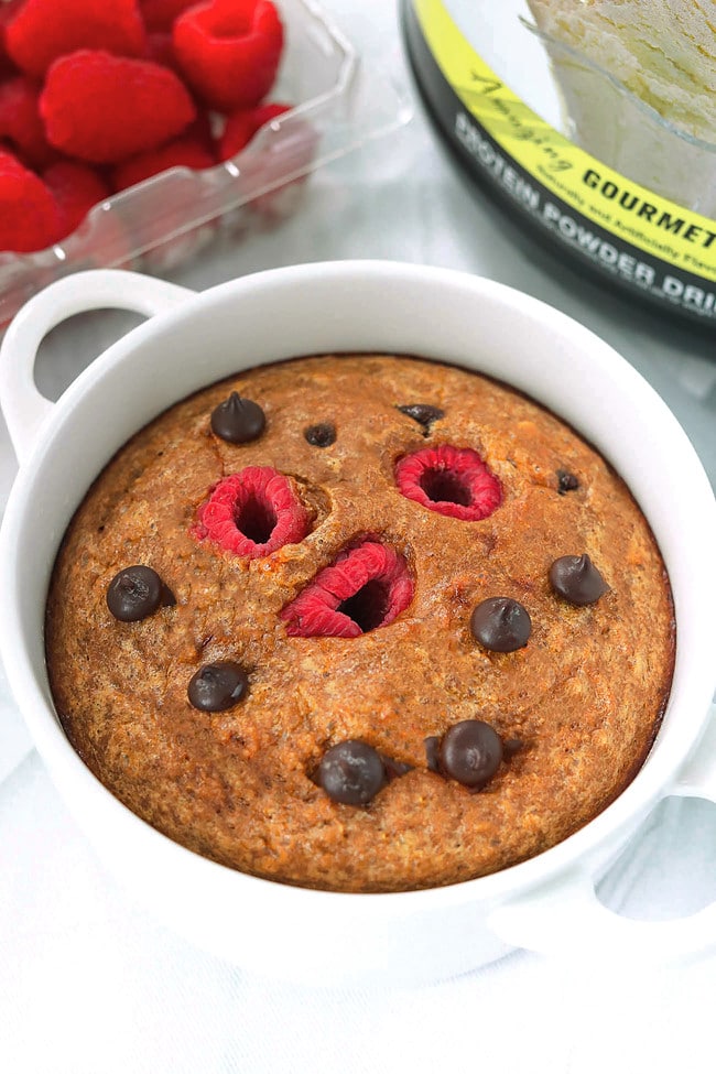 Front view of baked oats in a ramekin and protein powder tub and raspberries behind.