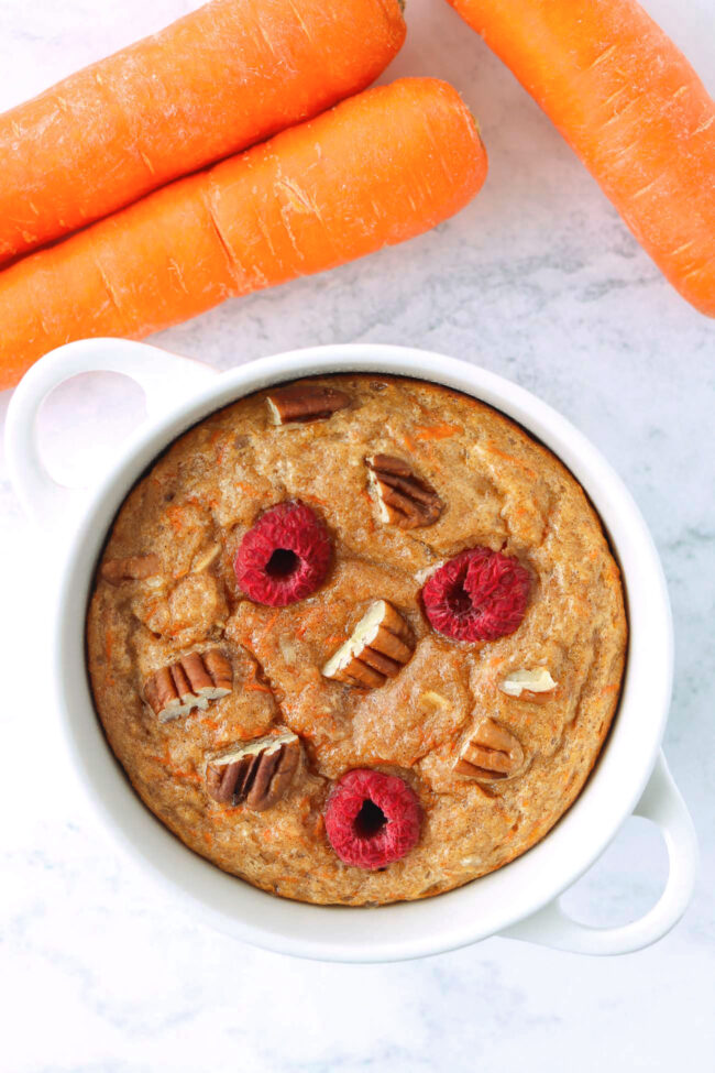 Carrot cake baked oats with pecans and raspberries in a ramekin and carrots on marble backdrop.