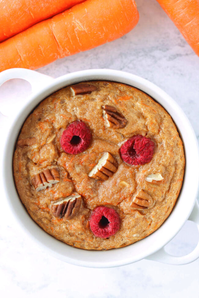 Carrot cake baked oats in a ramekin and carrots on marble backdrop.