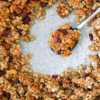 Granola on a spoon on a tray with granola clusters. Text overlay "Maple Tahini Granola", "Easy | Vegan | GF | DF" and "thatspicychick.com".