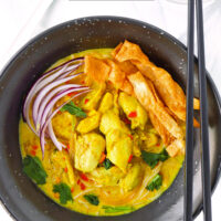 Chicken khao soi in bowl with sliced red onion, wonton strips and coriander and chopsticks. Text overlay "Easy Khao Soi", "Thai Coconut Curry Noodle Soup" and "thatspicychick.com".