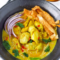 Front view of of chicken khao soi in bowl with sliced red onion, wonton strips and coriander. Text overlay "Easy Khao Soi", "Thai Coconut Curry Noodle Soup" and "thatspicychick.com".