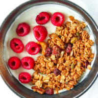 Bowl with Greek yogurt, granola, and fresh raspberries. Text overlay "Peanut Butter Granola", "Easy | Vegan | Gluten-free" and "thatspicychick.com".
