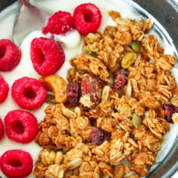 Bowl with spoon, granola and raspberries on Greek yogurt. Text overlay "Peanut Butter Granola", "High Protein | Gluten-free" and "thatspicychick.com".