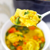 Spoon with a bite of tortellini soup. Text overlay "Thai Yellow Curry Tortellini Soup" and "thatspicychick.com".