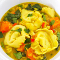 Closeup front view of curry tortellini soup in a bowl. Text overlay "Thai Yellow Curry Tortellini Soup" and "thatspicychick.com".