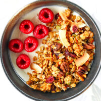 Top view of homemade granola in a bowl with raspberries and yogurt. Text overlay "Carrot Cake Granola", "Gluten-free | Vegan" and "thatspicychick.com"