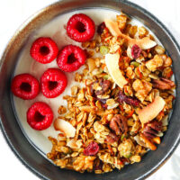 Close-up top view of homemade granola in a bowl with yogurt and raspberries. Text overlay "Carrot Cake Granola", "Healthy | Gluten-free | Vegan" and "thatspicychick.com"
