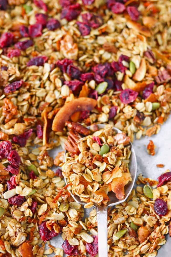 Close-up of large spoon in a parchment lined baking sheet pan with homemade carrot cake granola.