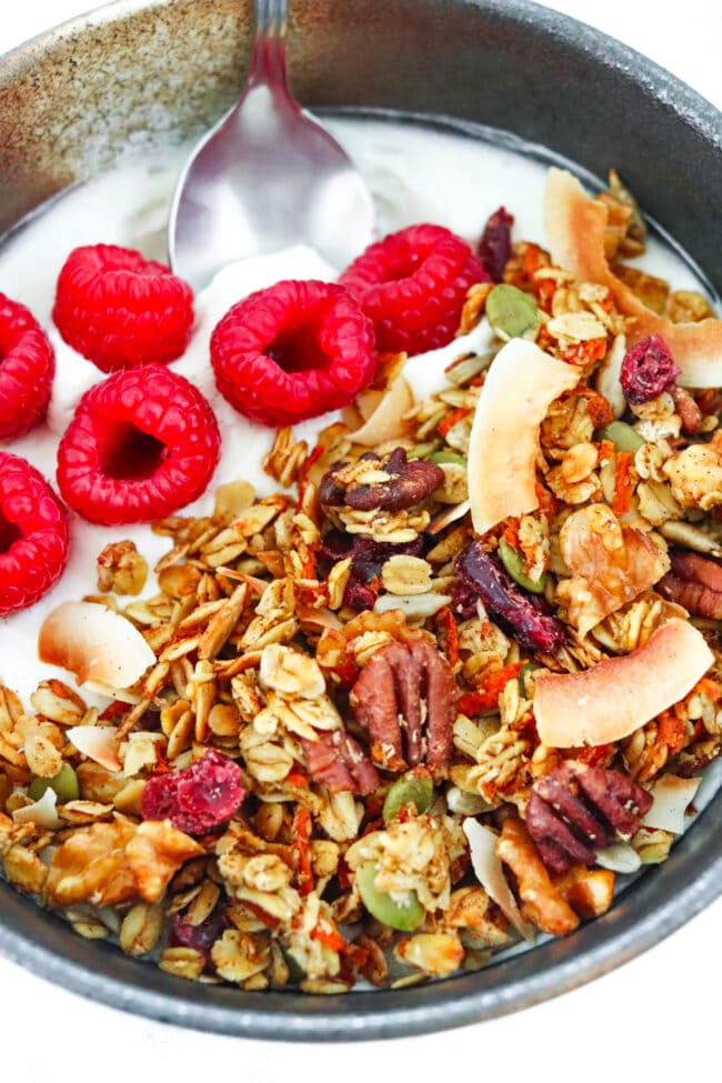 Close-up front view of bowl with yogurt, carrot cake granola, raspberries and a spoon.