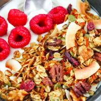 Close-up front view of granola on yogurt in a bowl with raspberries and a spoon. Text overlay "Carrot Cake Granola", "Healthy | Gluten-free" and "thatspicychick.com"