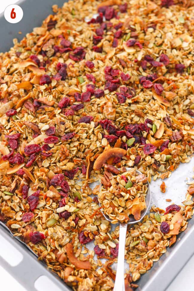 Dried cranberries mixed into carrot cake granola on a baking sheet with a large spoon.