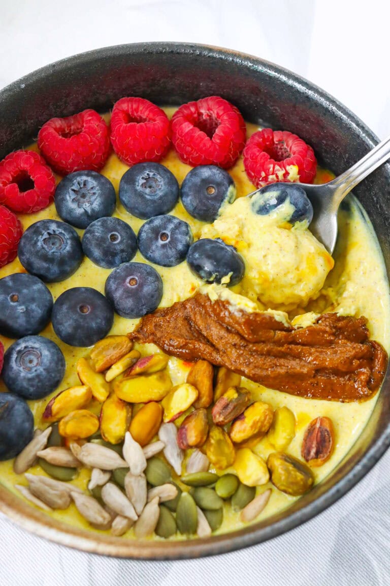 Closeup front view of spoon in bowl of golden milk overnight oats with berries, seeds, pistachios and pistachio butter.